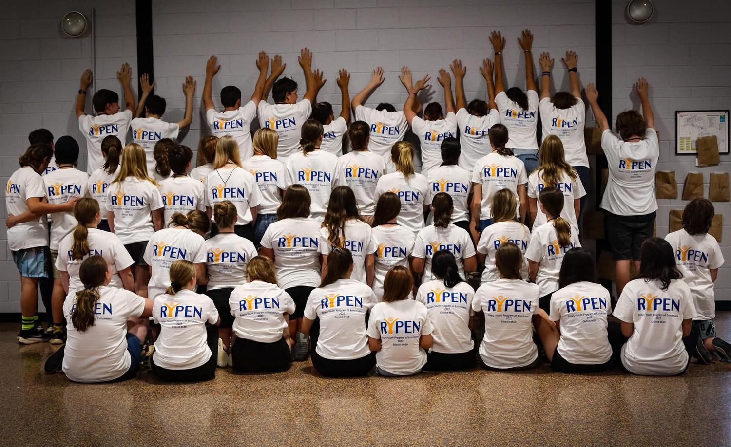 The 2022 group in white shirts, hands raised against the hall wall