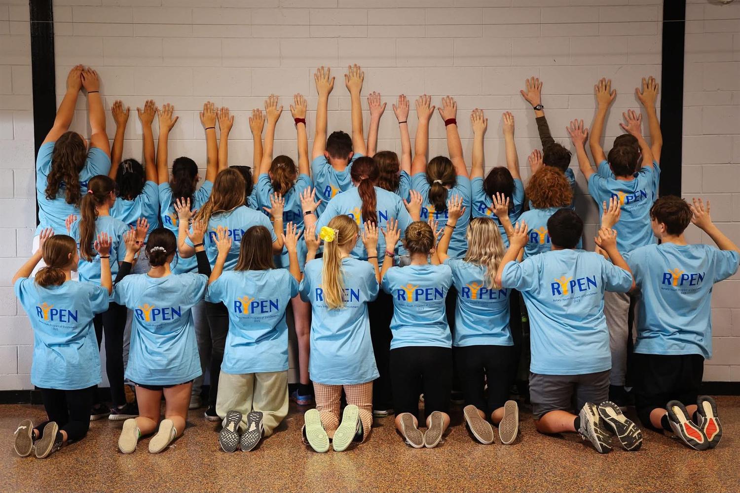 The 2023 group in blue shirts, hands raised against the hall wall
